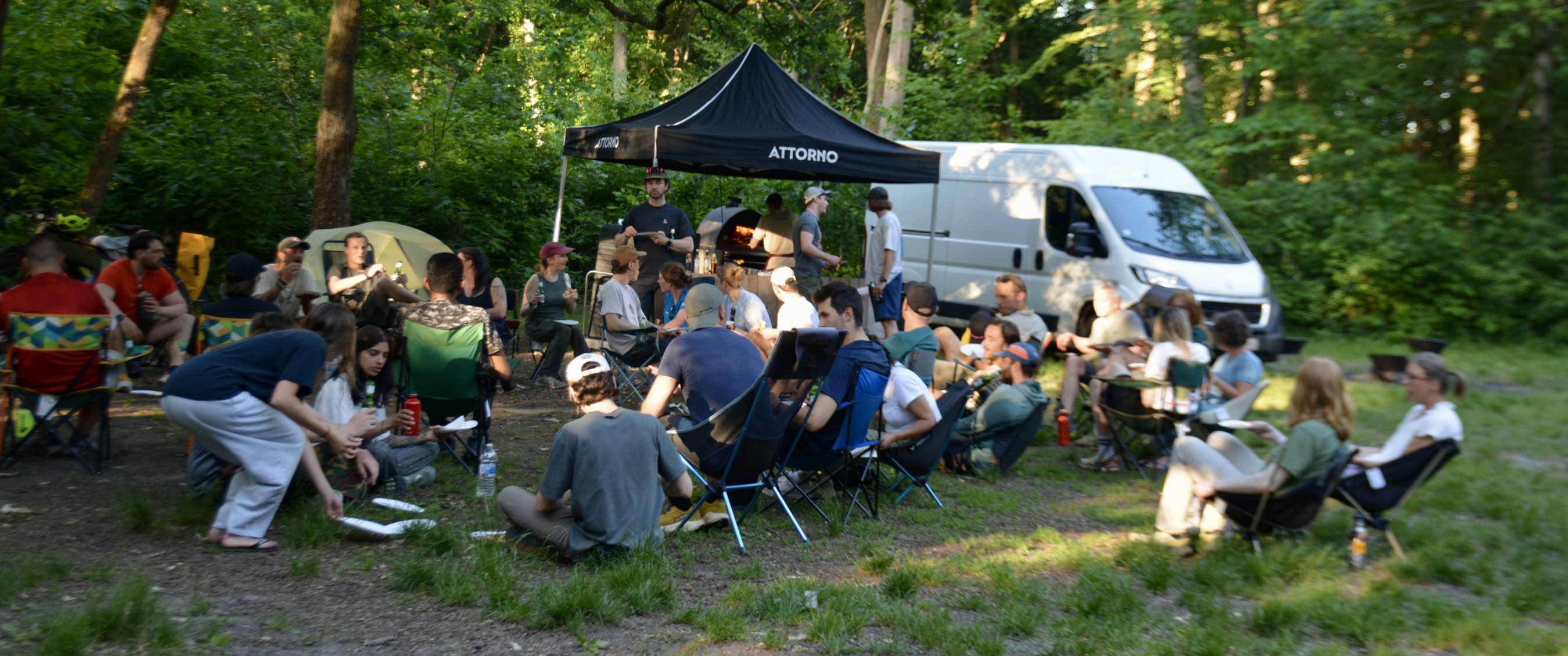 Groep mensen zit in stoelen en op de grond bij een zwarte ATTORNO-partytent in het bos.